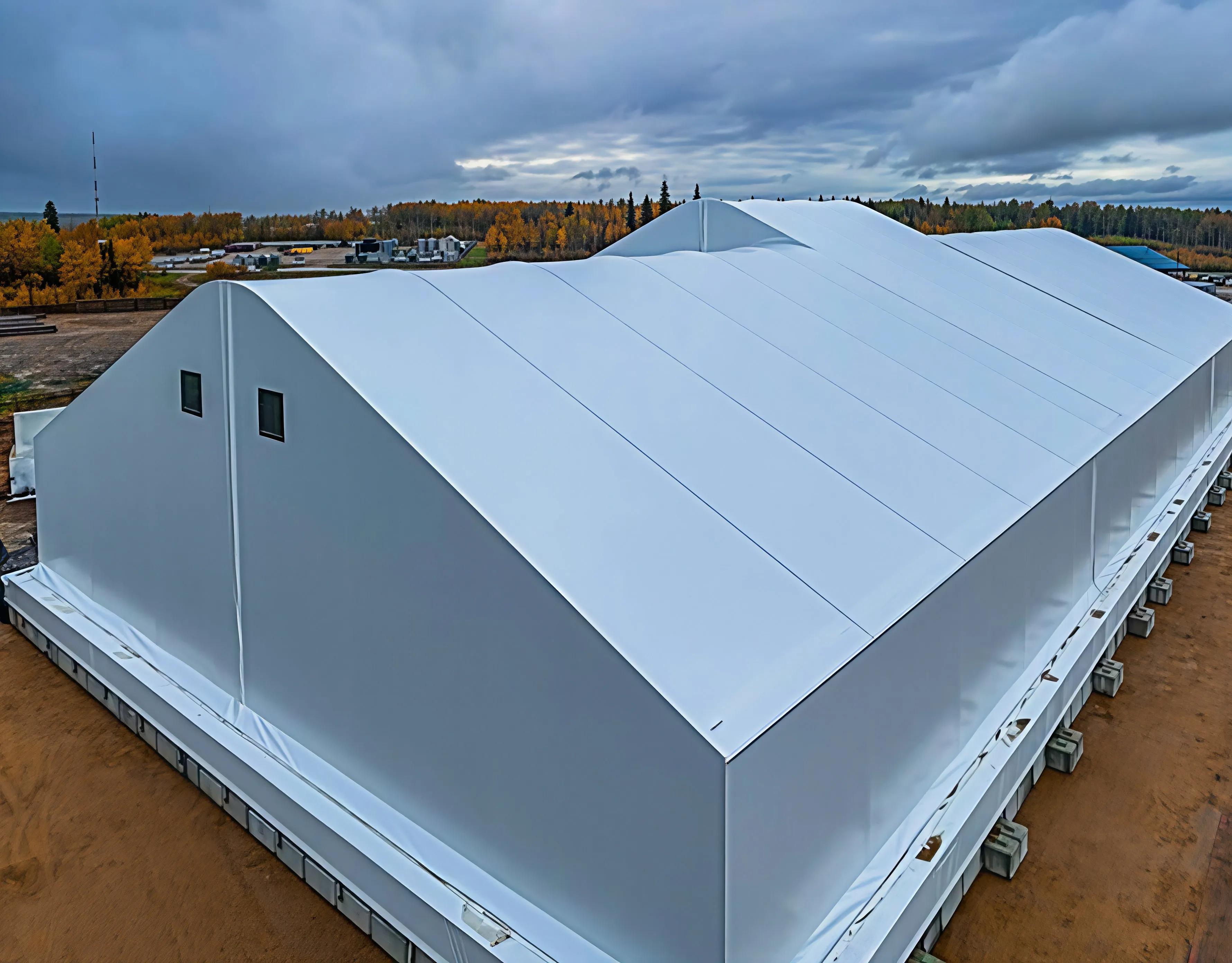 Aerial view of a white clear-span fabric building on a prepared site, with autumn boreal forest and overcast sky behind